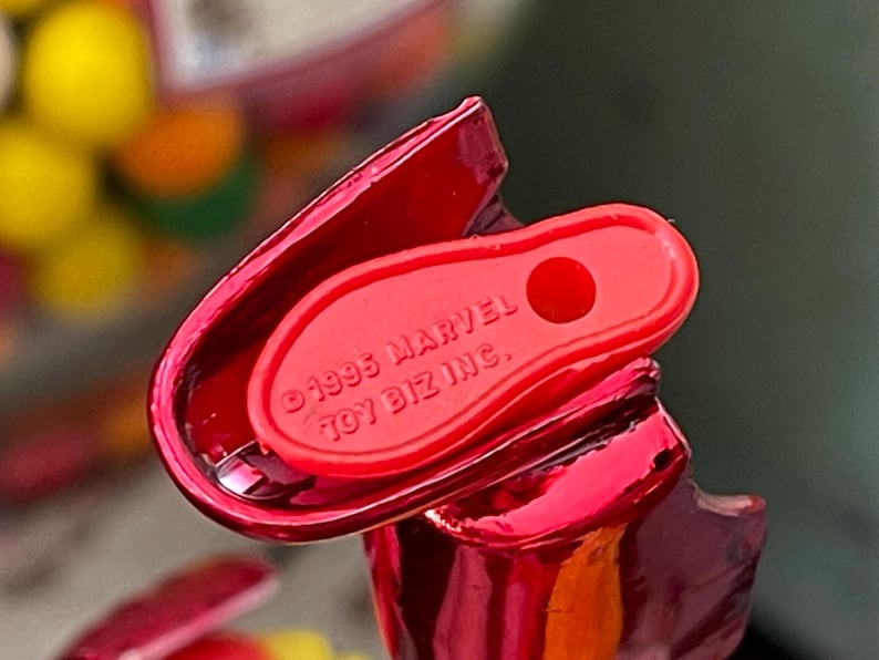 Może przedstawiać: Zbliżenie czerwonego plastikowego elementu zabawki z błyszczącym wykończeniem. Element ma zakrzywiony kształt i małą, płaską sekcję z tekstem "&copy;1995 MARVEL TOY BIZ INC.". Tło jest rozmyte.