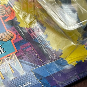 May include: Close-up of a plastic-wrapped action figure with a black and gray color scheme. The figure is partially visible and appears to be a superhero. The packaging features a yellow background with a cityscape and the word "WYTHE" in large, white letters.