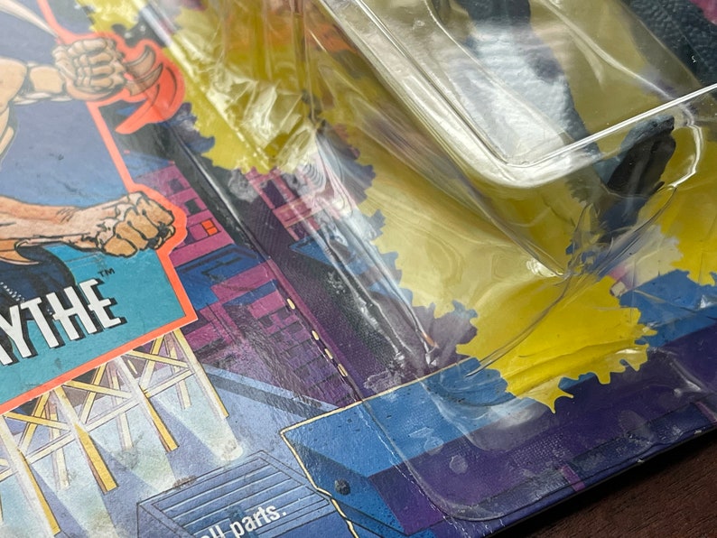 May include: Close-up of a plastic-wrapped action figure with a black and gray color scheme. The figure is partially visible and appears to be a superhero. The packaging features a yellow background with a cityscape and the word "WYTHE" in large, white letters.