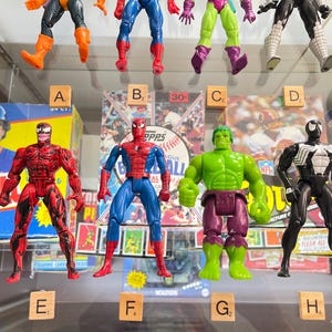 May include: A collection of action figures featuring comic book characters. The figures include Spider-Man, Hulk, Venom, and Carnage. The figures are displayed in front of baseball card boxes and a baseball-themed box.