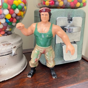 May include: A vintage action figure of a muscular man in a green tank top, camouflage pants, and black boots. The figure is posed in a fighting stance. The figure is next to a gumball machine.