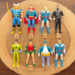 May include: A set of 8 action figures, including Superman, Robin, The Penguin, and The Riddler. The figures are arranged in a circle on a wooden surface. Each figure is labeled with a letter from A to H.