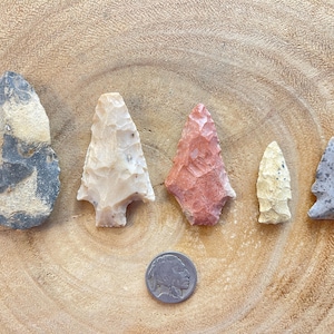 May include: Five arrowheads of different colors and shapes, including gray, white, red, and brown, are arranged on a wooden surface. A silver coin is in the center of the image.