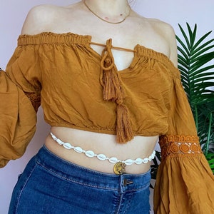 May include: A woman wearing a brown off-the-shoulder top with a tassel tie and a white shell bead belt. The top has long sleeves with lace trim. She is wearing blue jeans.
