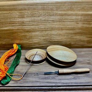 Może przedstawiać: Zestaw narzędzi do szycia na drewnianej powierzchni. Na zdjęciu znajdują się dwa drewniane uchwyty na nici, igła, szpikulec do prucia oraz pomarańczowa i zielona nić do haftowania. Drewniane przedmioty mają naturalne usłojenie.