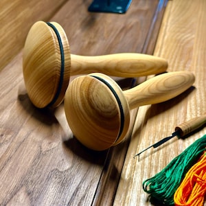 Może przedstawiać: Dwa drewniane narzędzia do szydełkowania dywanów z okrągłą główką i uchwytem, ​​obok igły i kolorowej nici do haftowania. Narzędzia mają jasne wykończenie słojów drewna z czarnym paskiem wokół główki. Nić jest zielona i pomarańczowa.