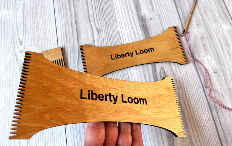May include: Two wooden weaving looms with the text "Liberty Loom" engraved on them. The looms are made of light brown wood and have a curved shape. A pink yarn is threaded through the loom on the right.