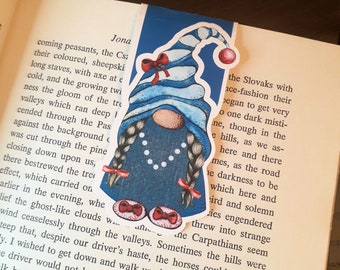 Op de afbeelding: Een blauwe gnome-bladwijzer met rode en witte accenten. De gnome draagt ​​een blauwe hoed met witte rand en een rode strik. De gnome heeft lange vlechten met rode strikken aan de uiteinden. De bladwijzer is versierd met witte stippen.