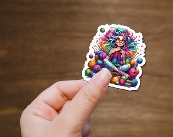 Op de afbeelding: Een kleurrijke sticker met een cartoonmeisje met regenbooghaar en een bril die breit terwijl ze op een stapel garenbollen zit.