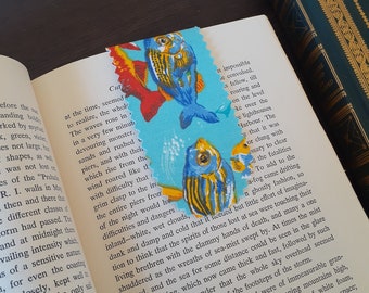 Op de afbeelding: Een blauwe en gele vis-bladwijzer met een turquoise achtergrond. De bladwijzer is boven en onder gescheurd. De tekst op de pagina luidt: "Cut at the time, seemed to realize, the whole. The waves rose in a very few minutes, devouring monster. Sands and rushed piers, and with them which rise from the wind roared like the with difficulty that grim clasp to the ire the entire piers from of the night would ha difficulties and dangers inland-white, wet clouds, dank and damp and cold that it liceued but little effort of imagina-tion to think that the spirits of those lost at sea were touching their living brethren with the clammy hands of death, and many a one shuddered as the wreaths of sea-mist swept by. At times the mist cleared, and the sea for some distance could be seen in the glare of the lightning, which now came thick and fast, followed by such sudden peals of thunder that the whole sky overhead seemed mmbling under the shock of the footsteps of the storm. of the scenes thus revealed were of immeasurable gran-thing interest-the sea, running mountains high, th wave mighty masses of white foam, th at and whirl away into space; of sail, running madly for wings of a storm-w search"