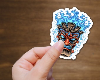 May include: A sticker featuring a wooden tiki mask with blue flames. The mask has red and blue details and a fierce expression.