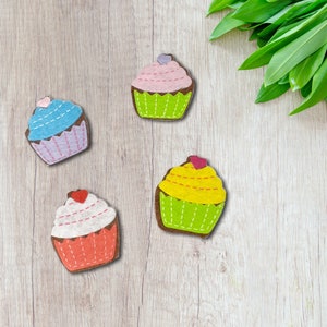 May include: Four felt cupcake decorations in different colors. The cupcakes are pink, blue, yellow, and red with white frosting. The cupcakes are on a light wood background.