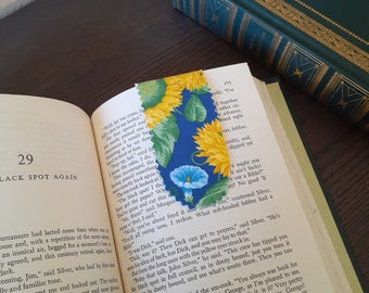 Op de afbeelding: Een stoffen bladwijzer met gele zonnebloemen en blauwe bloemen op een blauwe achtergrond, geplaatst in een open boek. Het boek is geopend op pagina 29. De bladwijzer is ongeveer 10 cm lang.