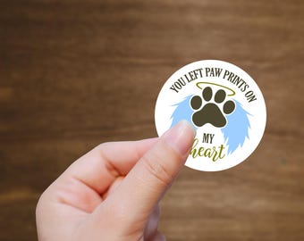 May include: White round sticker with the text "You left paw prints on my heart". A black paw print with a halo is in the center of the sticker. The paw print is surrounded by two blue angel wings.