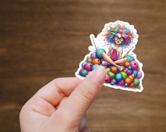 Op de afbeelding: Een kleurrijke sticker met een cartoonmeisje met regenbooghaar en een bril, zittend op een stapel garenbollen. Ze houdt breinaalden en een bol garen vast.