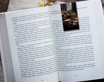 May include: A close-up of an open book with a bookmark. The bookmark is a stack of old books with a worn leather cover. The book is open to a page with text in Polish. The text on the page is about a warrior and a battle.