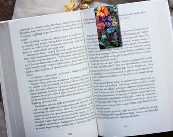 Op de afbeelding: Een close-up van een boek met een bladwijzer met een kleurrijk bloemenontwerp. De bladwijzer is gemaakt van papier en heeft een glanzende afwerking. Het boek is open op een pagina met tekst in een vreemde taal.