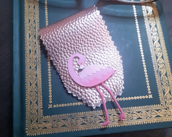 May include: A pink flamingo paper cutout on a gold leather bookmark. The bookmark is on a green book with a gold border.