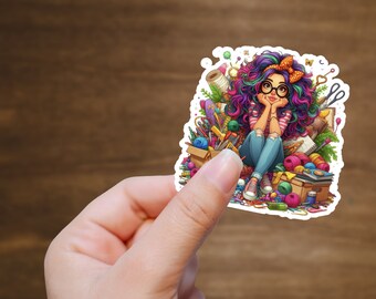 Op de afbeelding: Een kleurrijke sticker met een meisje met paars en roze haar dat een bril draagt en een rood-wit gestreept shirt. Ze zit op een stapel kleurrijke knutselspullen, waaronder garen, scharen en kwasten.