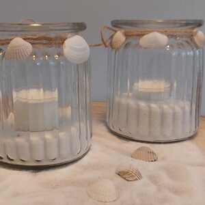 May include: Two clear glass candle holders with a coastal theme. Each jar is decorated with seashells and twine, and filled with white sand and a small candle. The jars are on a wooden surface with scattered seashells.