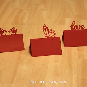 Puede incluir: Tres tarjetas rojas con recortes decorativos. Una presenta dos pájaros, otra una mariposa y la tercera la palabra "love". Las tarjetas se exhiben sobre una superficie de madera clara.