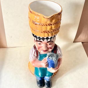 May include: A ceramic pitcher shaped like a man wearing a straw hat and holding a blue mug. The pitcher has a green apron and red and white striped shirt.