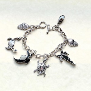 May include: A silver-toned charm bracelet featuring a toggle clasp and various marine-themed charms, including a sea turtle, a crab, a lobster, two spiral seashells, and a smooth oval stone, all attached to a thick, polished metal chain link.