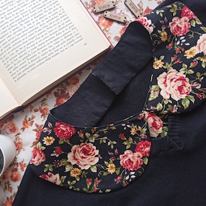 Puede incluir: Una camisa negra con cuello y un cuello con estampado floral. El cuello está hecho de tela negra con un estampado floral en tonos rosa, amarillo y verde.