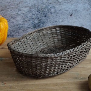 Peut inclure: Panier ovale en osier gris et marron. Le panier vide est posé sur une surface en bois, avec une courge jaune et des pommes de terre à proximité. La texture du panier est visible.