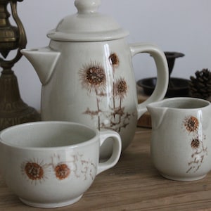 Puede incluir: Un juego de tazas de té de cerámica beige y una tetera con un diseño floral marrón. La tetera tiene una tapa y un asa. Las tazas de té tienen asas. El juego está sobre una mesa de madera.