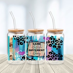 May include: Three glass drinking cups with bamboo lids and glass straws. Each cup features a colourful design with blue, teal, purple, and gold accents, and black leopard print. The centre cup has the text "SAME BITCH DIFFERENT DAY".