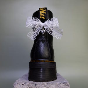 Könnte beinhalten: Ein schwarzer Lederstiefel mit dicker Profilsohle und gelben Nähten an der Basis. Eine weiße Spitzenschleife ist um die hintere Anziehschlaufe gebunden, an deren Mitte ein kleiner silberfarbener Herzanhänger befestigt ist.