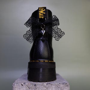 Könnte beinhalten: Ein schwarzer Lederstiefel mit dicker Plateausohle und gelben Nähten. An der Fersenschlaufe ist eine schwarze Spitzenschleife befestigt, die mit einem kleinen silberfarbenen Herzanhänger verziert ist. Die Fersenschlaufe ist mit gelbem Text versehen.