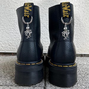 Könnte beinhalten: Ein Paar schwarze Lederstiefeletten mit genarbter Textur, dicken schwarzen Plateausohlen und gelben Nähten. Jeder Stiefel hat eine schwarze Fersenschlaufe mit gelbem Text und einen silberfarbenen Oktopus-Anhänger an einem Metallring.