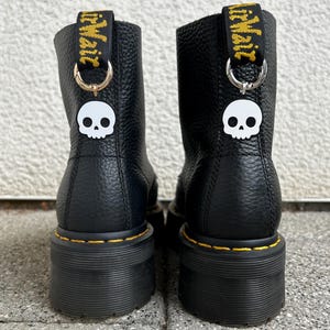 Könnte beinhalten: Ein Paar schwarze Lederstiefeletten mit genarbter Textur und dicken schwarzen Plateausohlen mit gelben Nähten. Jeder Stiefel hat eine schwarze Fersenschlaufe mit gelbem Text und einen silbernen Ring, an dem ein weißer Totenkopf-Anhänger befestigt ist.