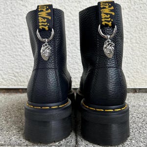 Könnte beinhalten: Ein Paar schwarze Lederstiefeletten mit genarbter Textur, ausgestattet mit dicken schwarzen Plateausohlen und gelben Nähten. Jeder Stiefel hat eine schwarze Fersenschlaufe mit gelbem Text und einen silbernen, anatomischen Herzanhänger am Ring.