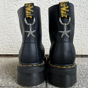 Könnte beinhalten: Ein Paar schwarze Lederstiefel mit genarbter Textur und dicken schwarzen Sohlen mit gelben Nähten. Jeder Stiefel hat eine schwarze Fersenschlaufe mit gelbem AirWair-Schriftzug, an der ein silberner Ring mit einem silbernen Seestern-Anhänger befestigt ist.