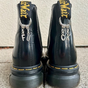 Könnte beinhalten: Paar schwarze Leder-Ankle Boots mit gelben Nähten und dicker Sohle. Jeder Stiefel hat eine gelb-schwarze Zuglasche mit dem Wort "AirWair". Silberne Charms buchstabieren "F U" und "C K" auf der Rückseite.
