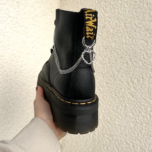 Könnte beinhalten: Schwarzer Plateau-Stiefel mit silberner Kette und Herzanhänger. Der Stiefel hat gelbe Nähte und eine gelb-schwarze Zuglasche mit dem Text "AirWair". Der Stiefel hat ein Obermaterial aus strukturiertem Leder.