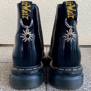 Könnte beinhalten: Schwarze Lederstiefel mit dicker Plateau-Sohle und gelber Naht. Die Stiefel haben einen Zuglasche mit dem Text "Dr. Martens" in Gelb. Jeder Stiefel hat einen silbernen Anhänger an der Lasche, der wie eine Sonne geformt ist.