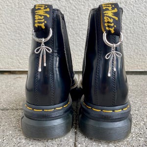 Könnte beinhalten: Schwarze Chelsea-Stiefel aus Leder mit einem gelben und schwarzen Schleifen-Zugband und einem silbernen Anhänger. Die Stiefel haben eine dicke schwarze Gummisohle.