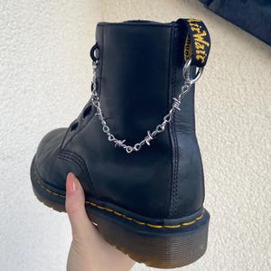 Könnte beinhalten: Schwarze Lederstiefel mit gelben Nähten und einer silbernen Kette mit Stacheldraht-Akzenten, die an der Oberseite des Stiefels befestigt sind.