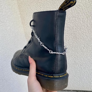 Könnte beinhalten: Schwarze Lederstiefel mit gelben Nähten und einer silbernen Kette mit Stacheldraht-Akzenten um den Knöchel.