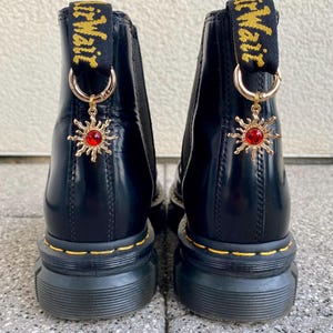 Könnte beinhalten: Schwarze Lederstiefel mit dicker Plateau-Sohle und einem goldenen Anhänger mit einem roten Juwel in Form einer Sonne. Die Stiefel haben einen gelben elastischen Zugriemen mit dem Aufdruck "Air Wair".