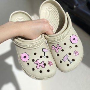 Könnte beinhalten: Ein Paar beigefarbene Clogs mit verschiedenen rosa und schwarzen Charms, darunter Blumen, Weingläser und Ballontiere.