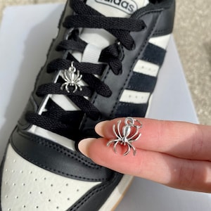 Könnte beinhalten: Schwarz-weiße Adidas Sneaker mit schwarzen Schnürsenkeln und silbernen Spinnen-Schuh-Anhängern.