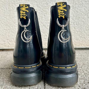 Könnte beinhalten: Schwarze Stiefeletten mit gelben Nähten und einer klobigen Sohle. Jeder Stiefel hat einen silbernen Halbmond-Anhänger, der an einer Schlaufe auf der Rückseite hängt. Die Stiefel haben ein glänzendes Finish und den Text "Air Wair" in Gelb.
