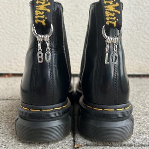 Könnte beinhalten: Paar schwarze Leder-Ankle Boots mit gelben Nähten und dicken Sohlen. Die Stiefel haben eine Zuglasche mit gelbem Text und silbernen Anhängern mit den Buchstaben "BO" und "LD".