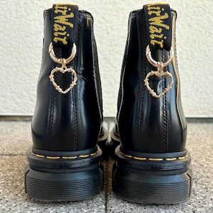 Könnte beinhalten: Schwarze Leder-Stiefeletten mit einer dicken Plateau-Sohle und einem goldenen Herz-Anhänger, der an der hinteren Lasche befestigt ist. Die Stiefeletten haben ein gelbes Stichdetail um die Sohle und der Text "Dr. Martens" ist auf der Lasche aufgedruckt.