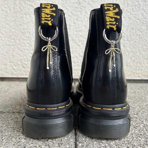 Könnte beinhalten: Schwarze Lederstiefel mit gelber Naht und einem goldenen Anhänger. Die Stiefel haben eine dicke Plateau-Sohle und einen Zuglaschen mit dem Text "Dr. Martens" in Gelb.